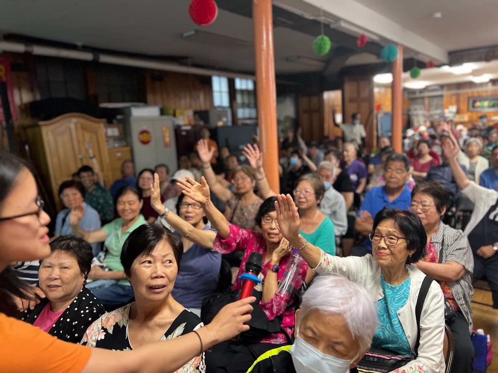 A group of older Asian people seated in chairs, with several women raising their hands in the center.